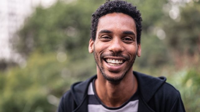 Man in black jacket with striped shirt smiling with a park in the background