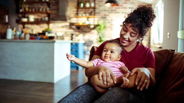 Woman holding 10-month-old baby girl while sitting on couch in kitchen